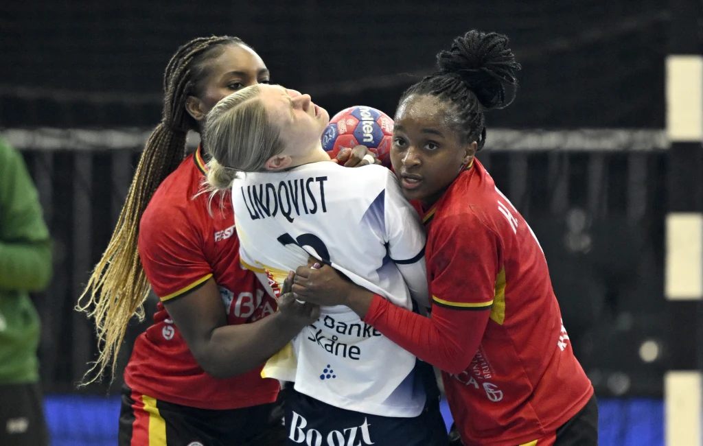 Emma Lindkvist zatrzymywana w meczu Angola - Szwecja Trzy zawodniczki piłki ręcznej podczas rywalizacji na boisku, dwie w czerwonych strojach intensywnie bronią, otaczając zawodniczkę w białej koszulce z piłką w rękach.