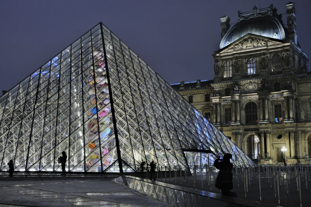 Szklana piramida Muzeum Luwru oświetlona nocą, tło stanowi zabytkowy budynek muzeum z klasyczną fasadą. Na pierwszym planie sylwetki ludzi, przyciemnione uliczne światła oraz odbicia na mokrym chodniku.