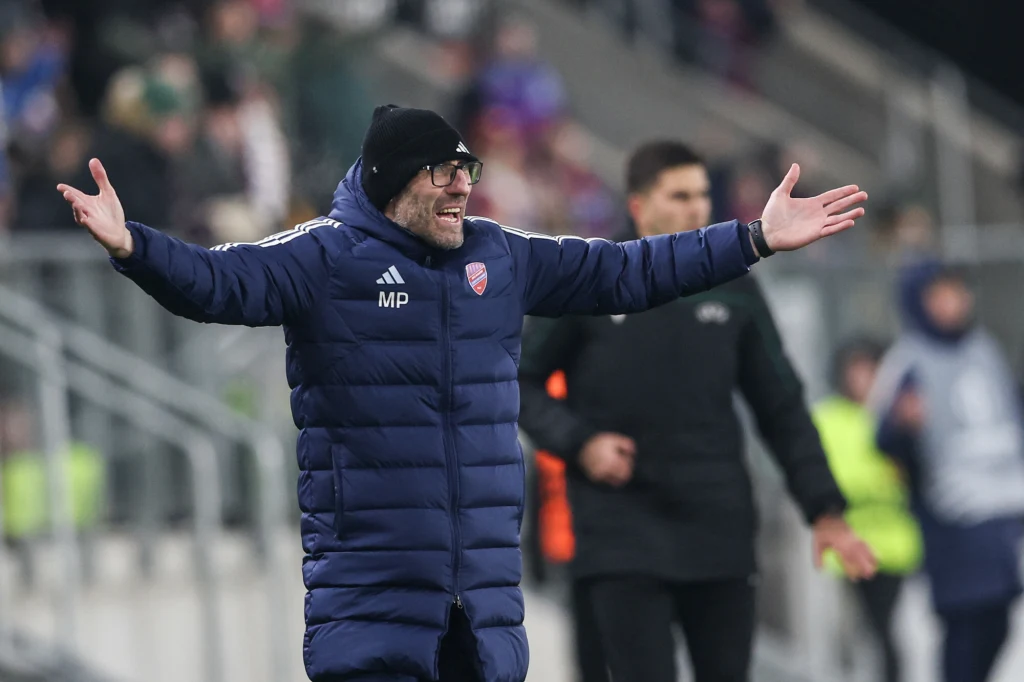 Marek Papszun Trener w kurtce zimowej z rozłożonymi rękoma, stojący na stadionie podczas meczu piłkarskiego, w tle rozmazane sylwetki innych osób.