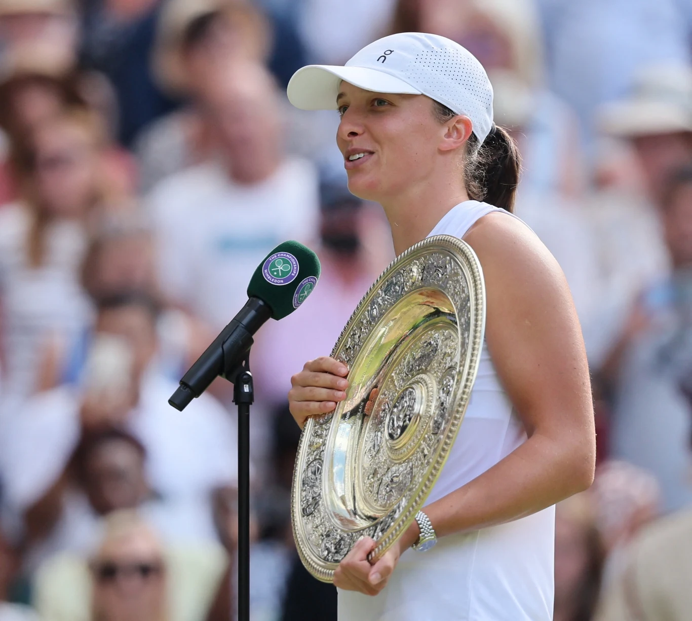 Kobieta ubrana w białą sportową odzież oraz czapkę z daszkiem trzyma srebrny, okrągły puchar, stojąc przy mikrofonie podczas ceremonii na tenisowym korcie. W tle widoczny tłum kibiców.