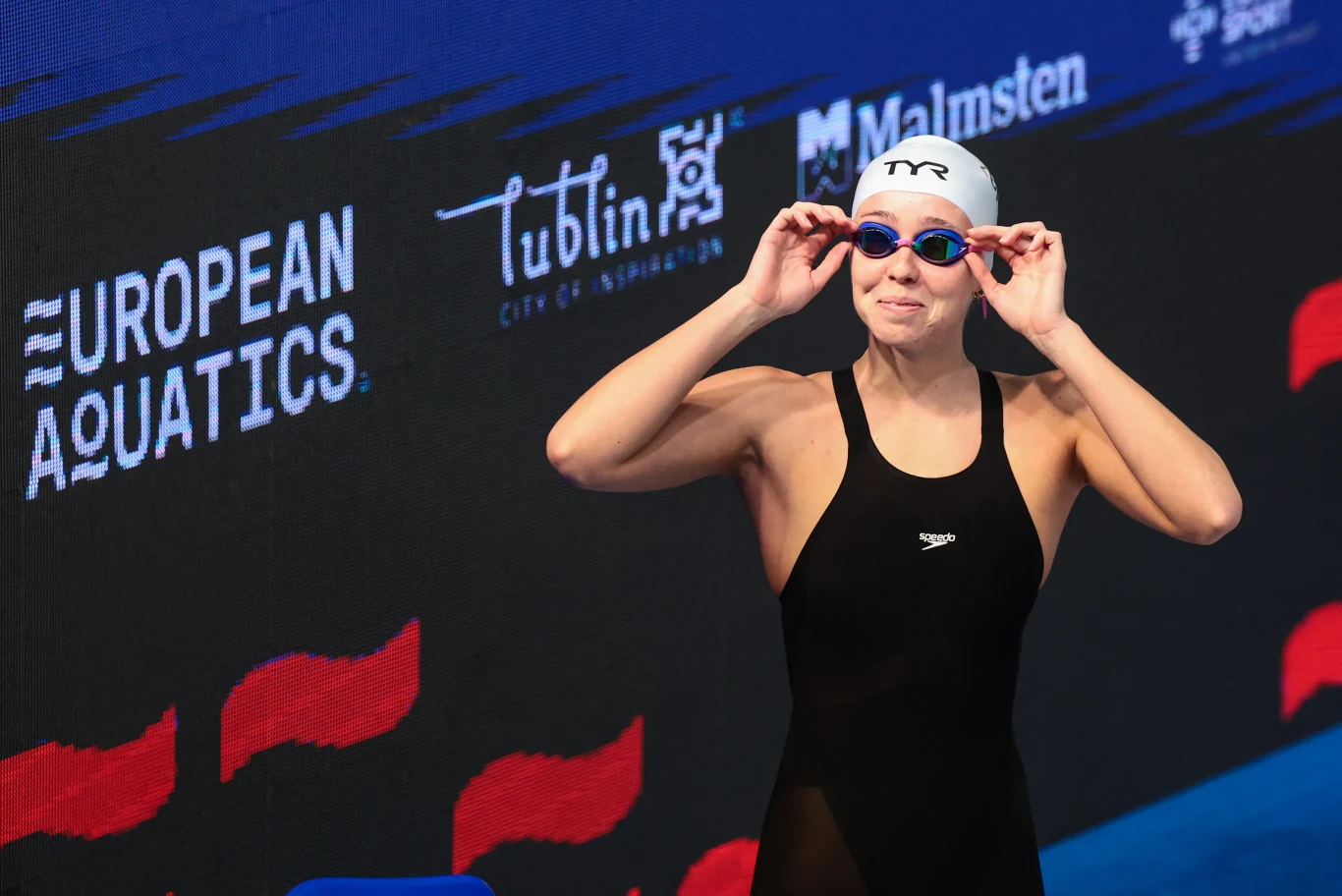 Zawodniczka w stroju pływackim oraz czepku zakłada okulary pływackie, stojąc przygotowana do startu na tle tablicy z logo European Aquatics oraz nazwami sponsorów.
