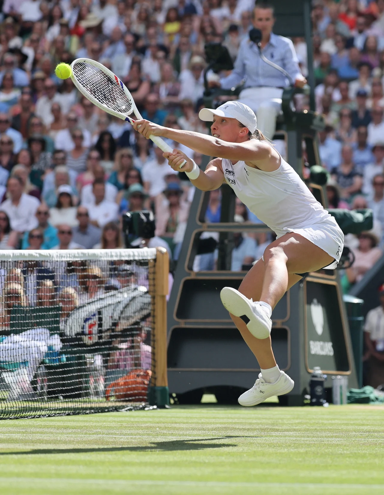Tenisistka w białym stroju wykonuje dynamiczny ruch przy siatce, próbując odbić piłkę rakietą podczas meczu na trawiastym korcie. W tle widać sędziego na podwyższeniu oraz licznych kibiców na trybunach.