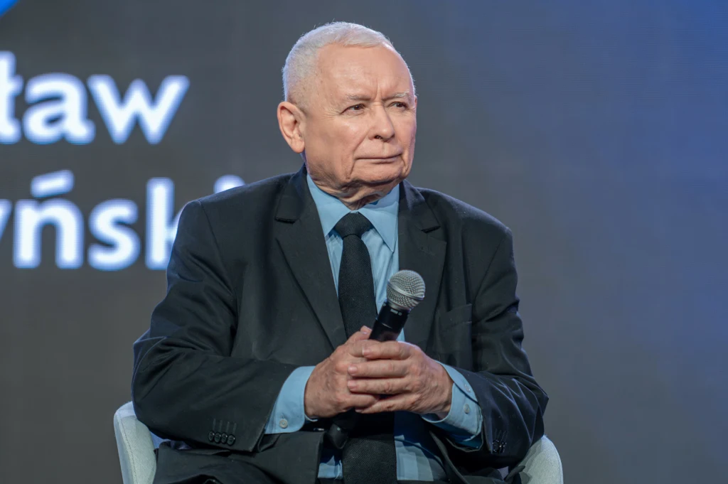 Jarosław Kaczyński zamieścił wpis. "Rzadko reaguję na kłamstwa" Starszy mężczyzna w garniturze siedzi na krześle, trzymając mikrofon w dłoniach, w tle rozmazany napis na granatowym tle.