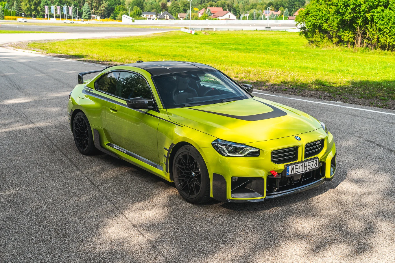 BMW M2 obecnie właściwie nie ma konkurentów na rynku Sportowy samochód BMW w jaskrawożółtym kolorze stoi zaparkowany przy drodze. Auto posiada sportowe felgi, aerodynamiczne dodatki oraz czarny dach i maskę. W tle widoczna jest zieleń i fragment zabudowy, całość oświetlona słonecznym światłem.