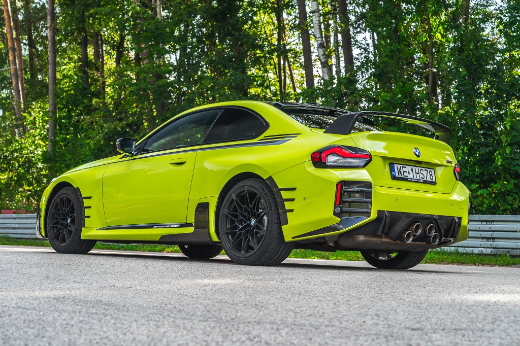 Sportowy samochód w jaskrawym, żółtym kolorze, zaparkowany na tle zielonych drzew, wyposażony w tylny spojler, czarne felgi oraz cztery końcówki wydechu.