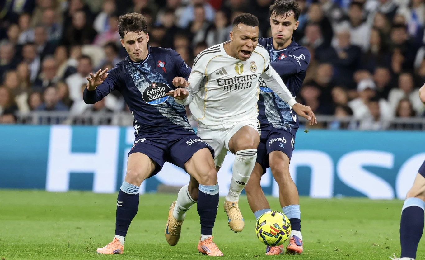 Kylian Mbappe wzięty "w kleszcze" przez defensorów Celty Vigo Dwaj zawodnicy w niebieskich strojach próbują odebrać piłkę piłkarzowi ubranemu na biało w trakcie dynamicznego zagrania na boisku, wokół widać publiczność na trybunach.