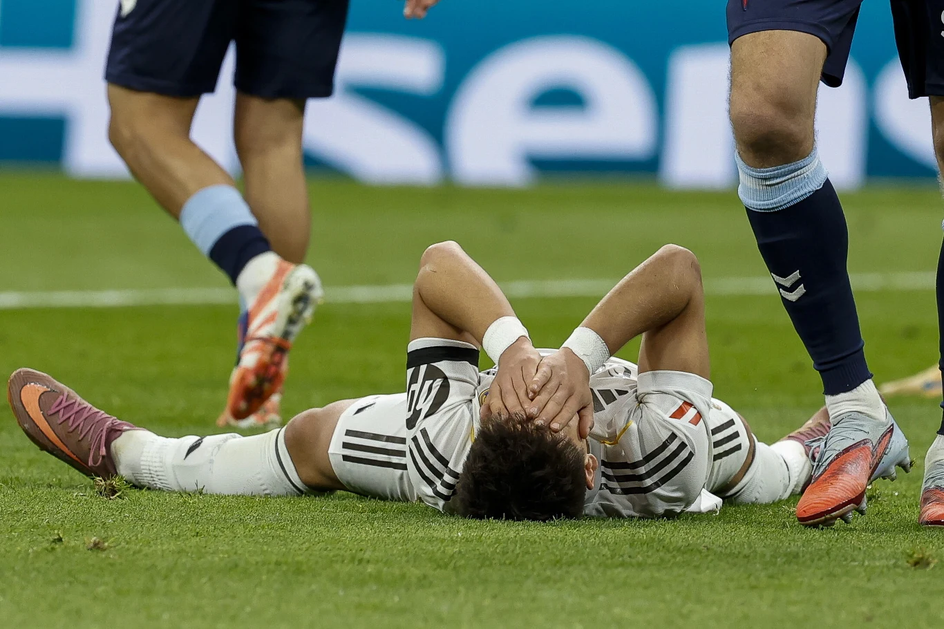 Wymowny obrazek. Arda Guler po kolejnym nieudanym natarciu Realu Madryt Zawodnik piłkarski w biało-czarnej koszulce leży na murawie, zakrywając twarz dłońmi. Wokół niego stoją inni piłkarze z drużyny przeciwnej.