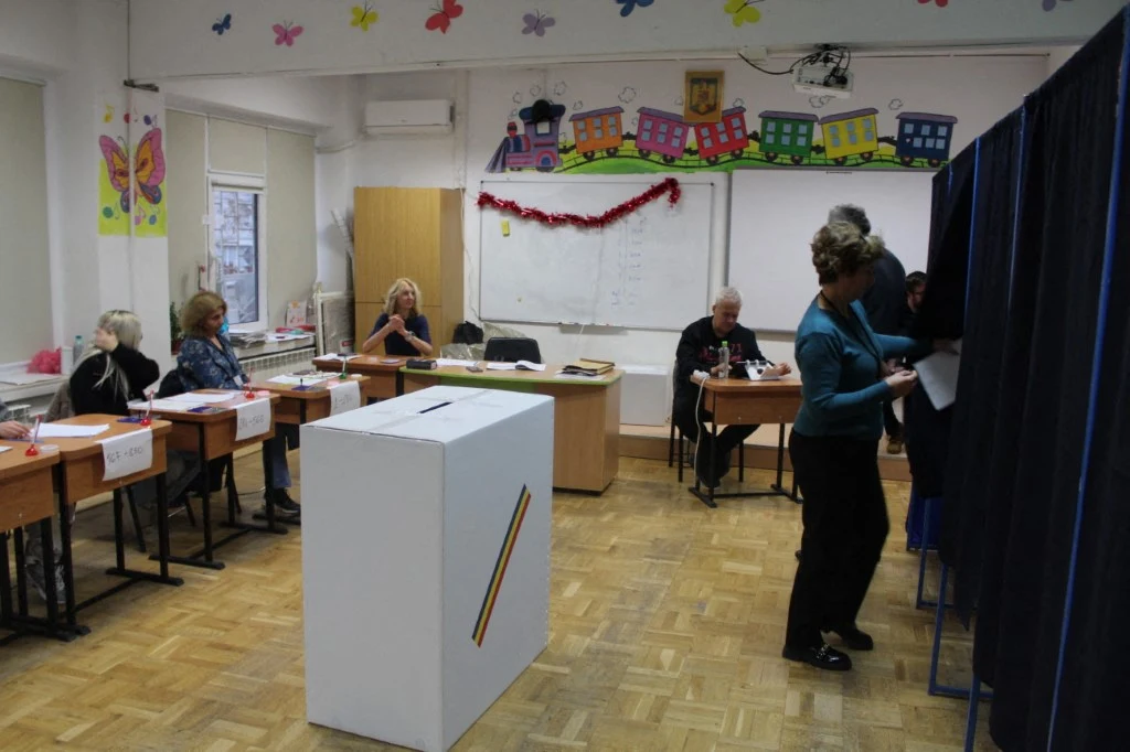 Wybory na burmistrza Bukaresztu. Liberał Ciprian Ciucu na prowadzeniu, pojawiają się częściowe wyniki Komisja wyborcza pracująca w udekorowanej klasie szkolnej, urny wyborcze na pierwszym planie oraz osoby głosujące oddające głosy za zasłoną; członkowie komisji siedzą przy stole obserwując przebieg wyborów.