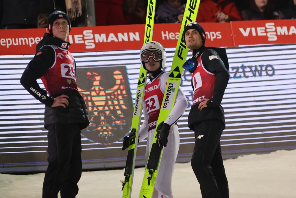 Od lewej: Paweł Wąsek. Kacper Tomasiak i Maciej Kot Trzech mężczyzn w strojach sportowych do skoków narciarskich stoi obok siebie na śniegu, jeden z nich trzyma narty Fischer. W tle znajduje się elektroniczna tablica z napisem Viessmann i godłem, widoczna publiczność.