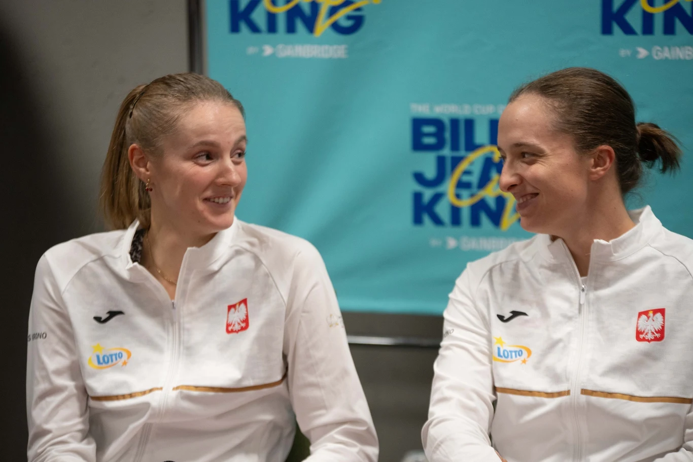 Martyna Kubka i Iga Świątek Dwie kobiety w białych sportowych kurtkach z polskim godłem i logotypami partnerów rozmawiają ze sobą, obie uśmiechnięte. W tle widoczny baner promocyjny z napisem Billie Jean King Cup.