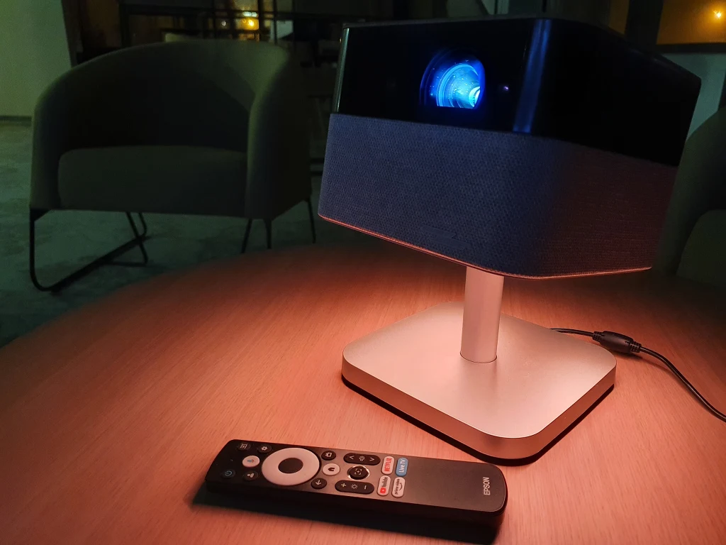Epson Flex EF-72 Projektor multimedialny z niebieskim światłem emitowanym z obiektywu ustawiony na stoliku, obok pilot zdalnego sterowania, w tle delikatnie oświetlone pomieszczenie z fotelami.