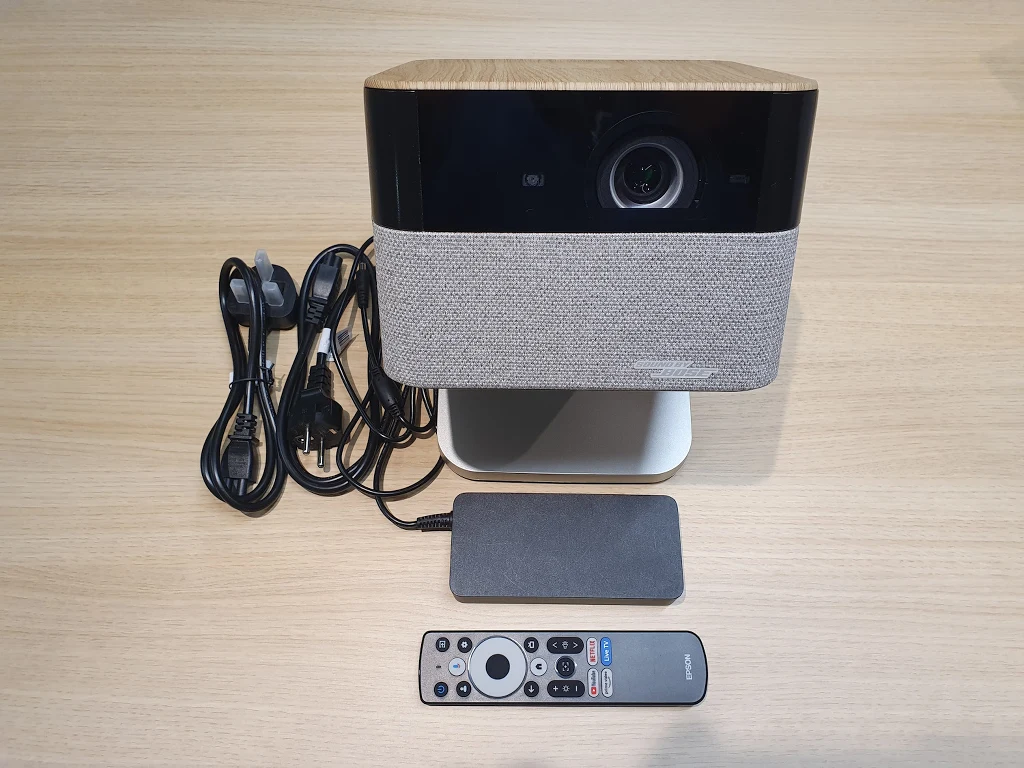 Epson Flex EF-72 Projektor multimedialny z obudową w kolorze drewna i szarości ustawiony na jasnej powierzchni, obok leżą dwa kable zasilające, zasilacz oraz pilot z przyciskami do sterowania.