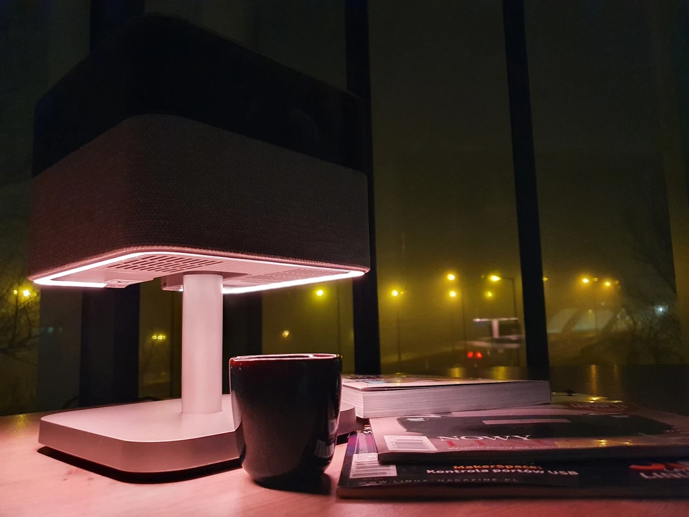 Nowoczesna lampa biurkowa świecąca ciepłym światłem, stojąca na blacie obok czarnego kubka oraz kilku rozłożonych czasopism i książek. W tle duże okno z widokiem na nocną, lekko zamgloną ulicę oświetloną żółtymi lampami ulicznymi.