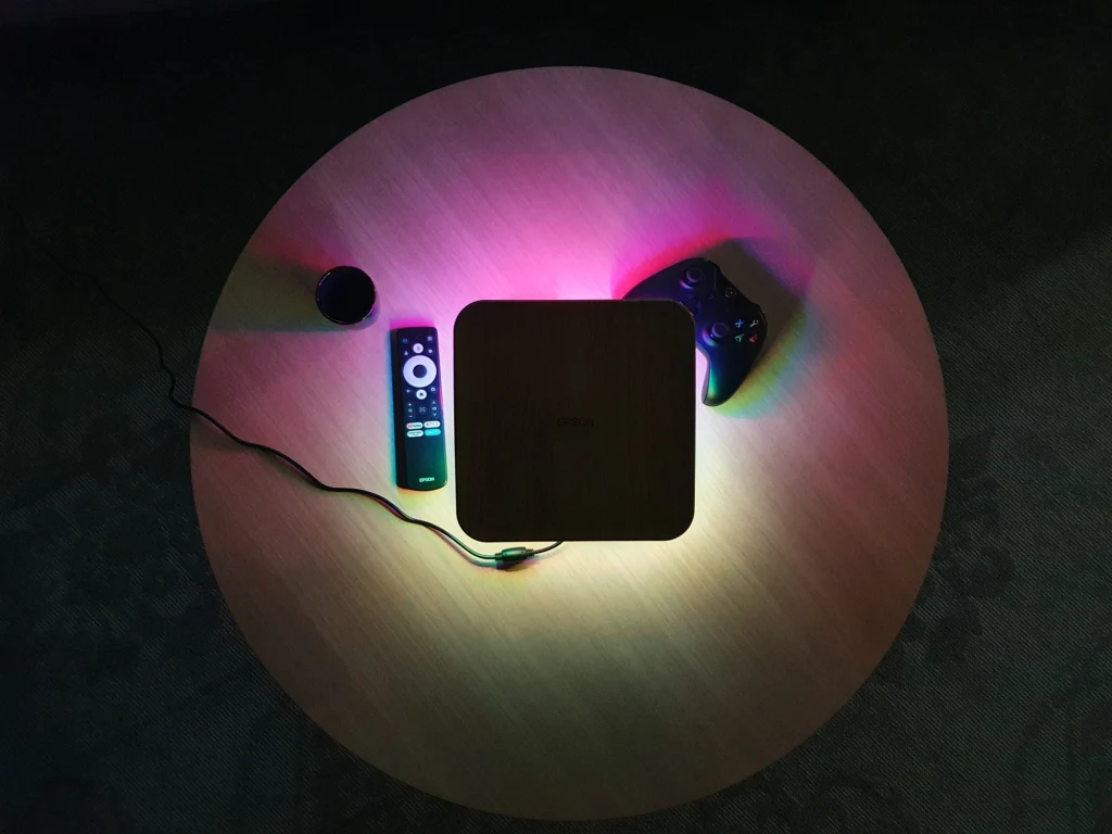 Epson Flex EF-72 Kwadratowa, czarna przystawka multimedialna ustawiona na środku okrągłego stołu, oświetlona różnokolorowym światłem LED. Obok przystawki leżą pilot, kontroler do gier oraz niewielki okrągły obiekt. Wszystkie elementy tworzą spójną, nowoczesną kompozycję.