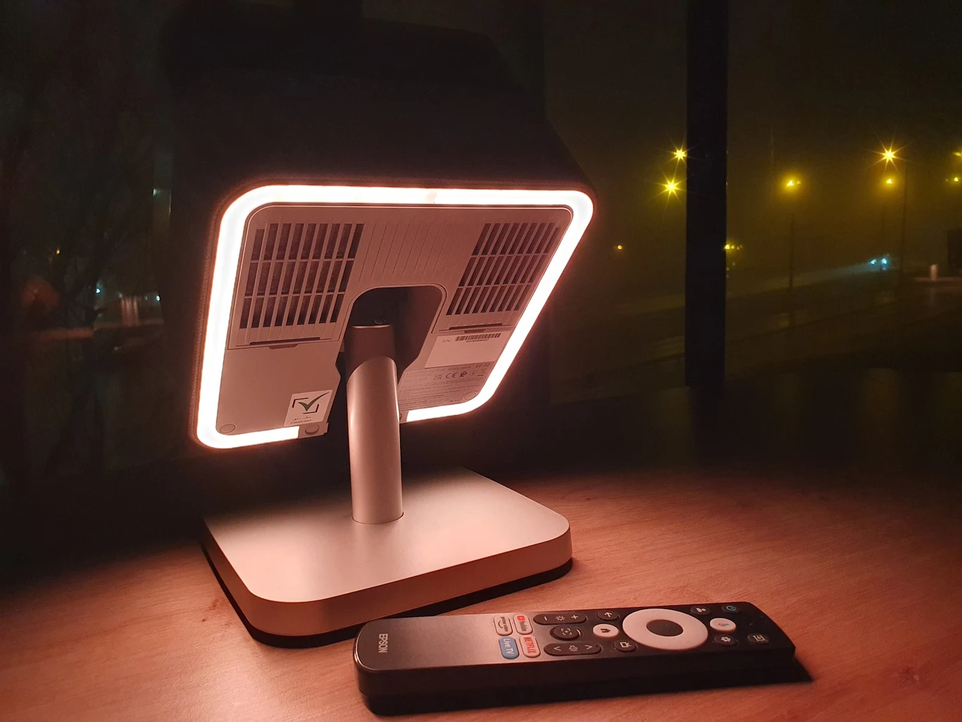 Nowoczesna lampka biurkowa oświetlająca blat, obok leży pilot zdalnego sterowania. W tle ciemne okno z widocznymi światłami ulicznymi.
