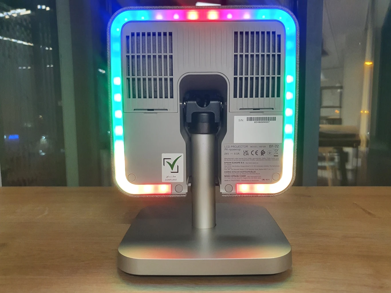 Tył nowoczesnego monitora lub urządzenia elektronicznego z aluminiową podstawą, podświetleniem LED RGB wokół obudowy oraz widocznymi wentylatorami chłodzącymi, ustawionego na drewnianym blacie przy oknie.