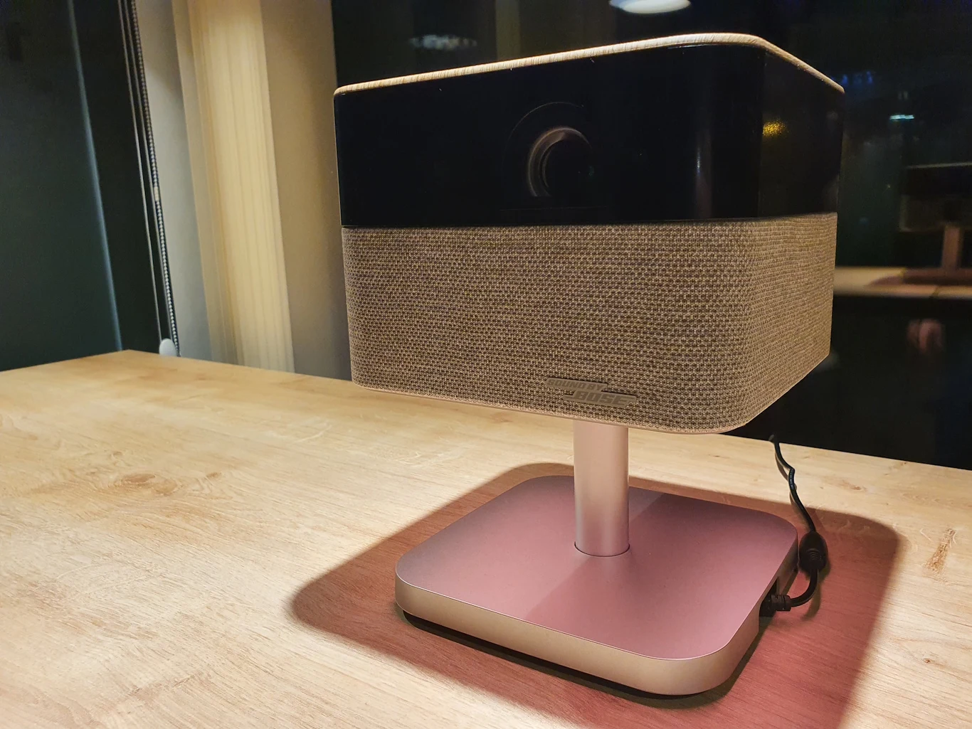 Nowoczesny projektor ustawiony na drewnianym stole, urządzenie o kwadratowej podstawie, z obudową wykończoną materiałem i przewodem zasilającym widocznym z boku.