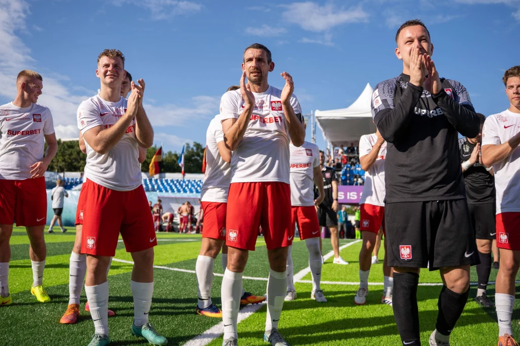 Reprezentacja Polski w socca Grupa piłkarzy ubrana w biało-czerwone stroje stoi na boisku, klaska w ręce i cieszy się po zakończeniu meczu, w tle widoczne są trybuny oraz niebieskie niebo.