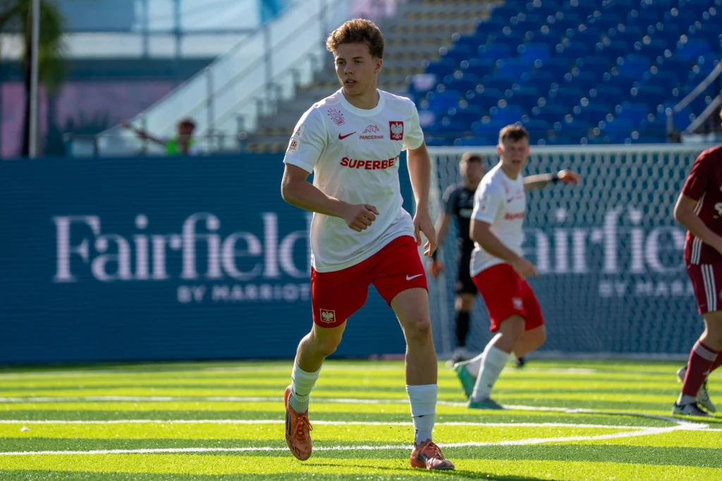 Jan Grzybowski Młody piłkarz w białej koszulce z logo Polski i czerwonych spodenkach biegnie po zielonej murawie, w tle widoczni inni zawodnicy oraz trybuny stadionu.