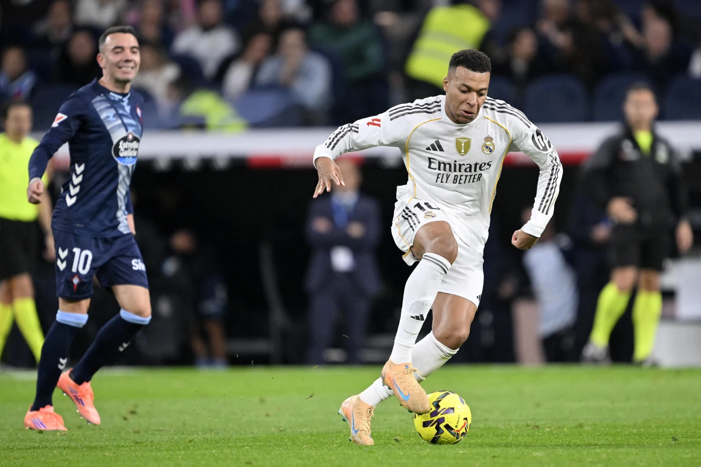 Piłkarz w stroju Realu Madryt prowadzi piłkę nożną podczas meczu, a obok niego biegnie zawodnik drużyny przeciwnej w niebieskim stroju; w tle widownia i sędziowie obserwują wydarzenia na boisku.