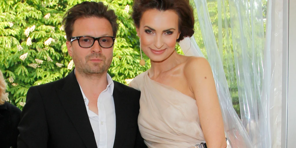 Agnieszka Macią z mężem, Robertem Wolańskim Para elegancko ubranych osób pozująca razem przy oknie z białą zasłoną, w tle zielone drzewa ogrodu. Kobieta w jasnej, asymetrycznej sukni wieczorowej uśmiecha się, mężczyzna w czarnym garniturze i białej koszuli ma okulary.