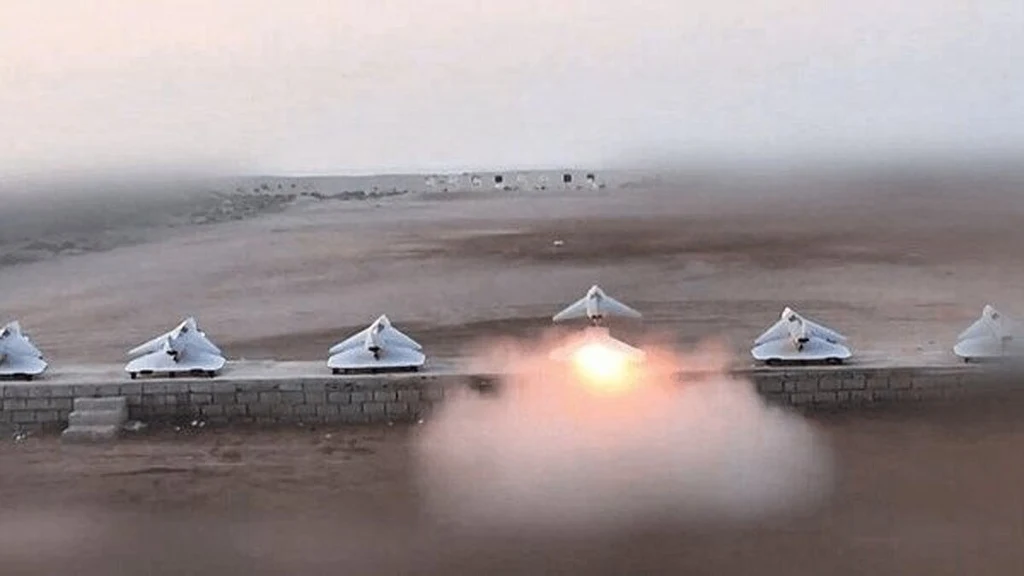 Rząd równo ustawionych na ziemi dronów, z których jeden startuje, emitując ogień i dym, pustynny krajobraz w tle.