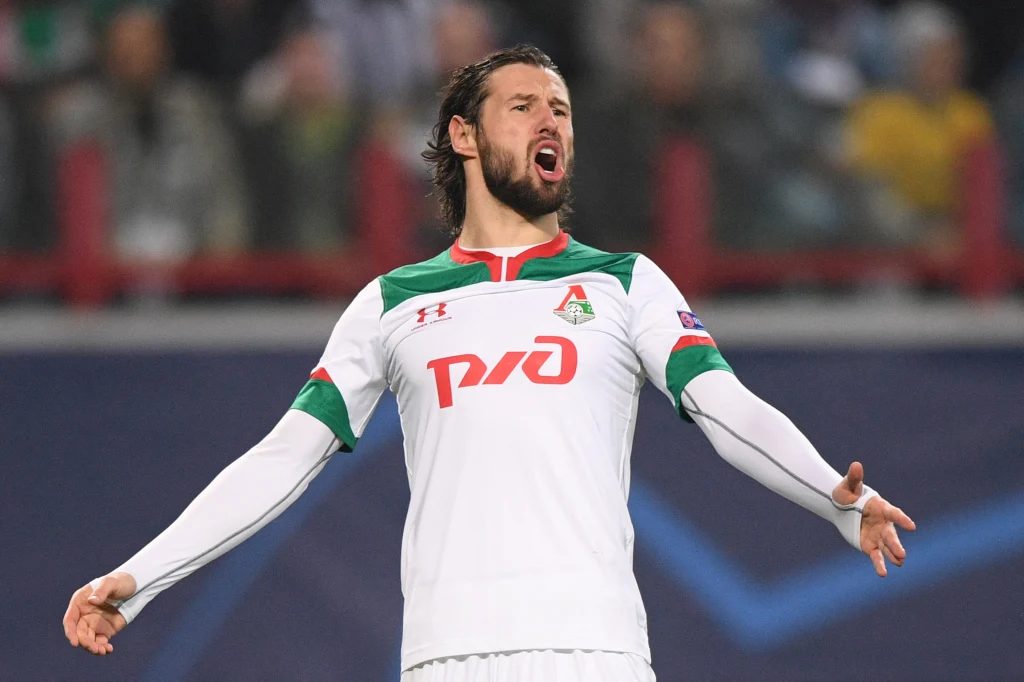 Grzegorz Krychowiak w barwach Łokomotiwu Moskwa Piłkarz w białym stroju z zielono-czerwonymi akcentami na boisku, z rozpostartymi ramionami i otwartymi ustami w geście wyrażającym emocje. W tle rozmyta publiczność stadionu.