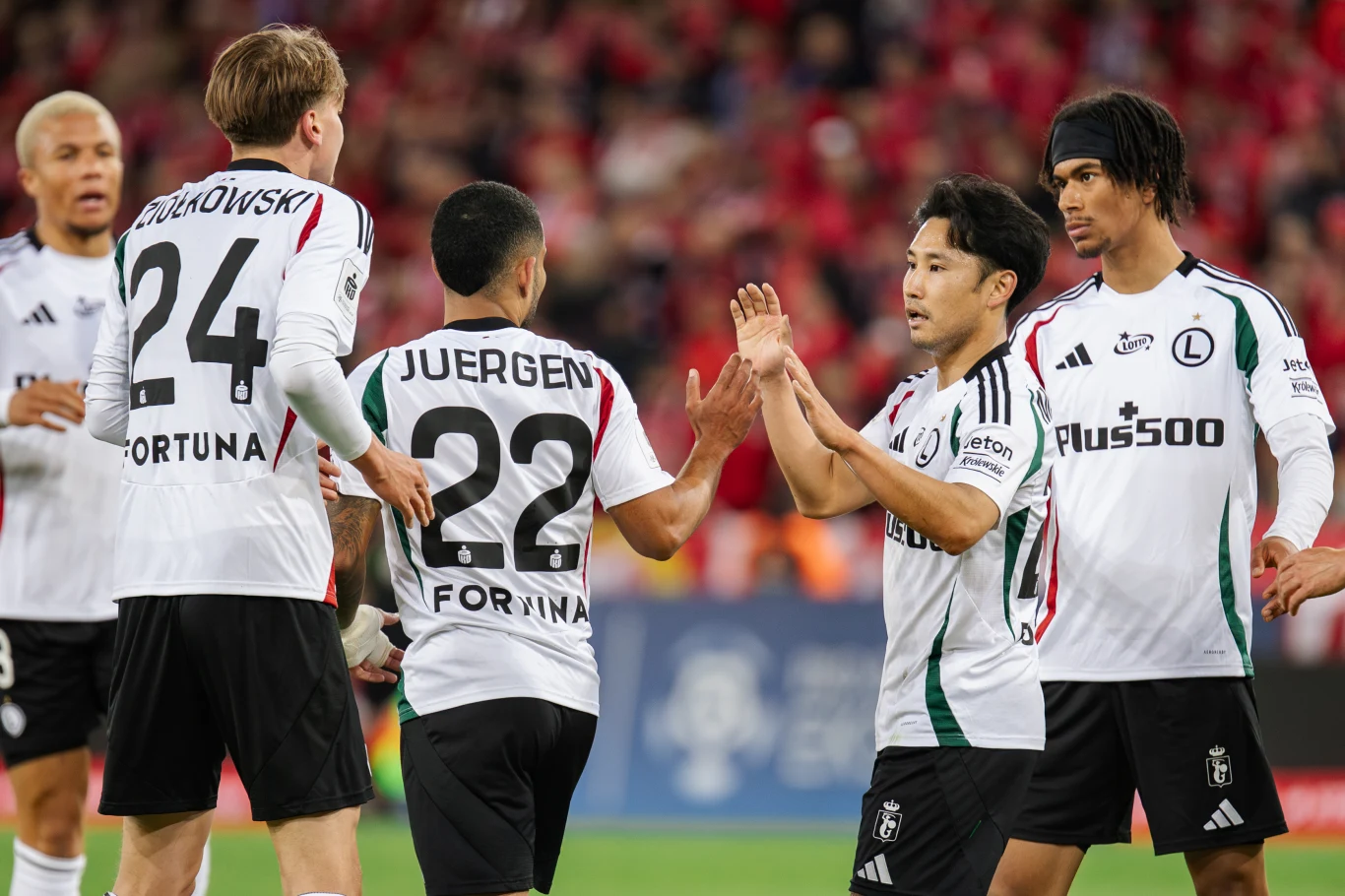 Ryoya Morishita (drugi z prawej) wręcz uwielbia Polskę Grupa piłkarzy Legii Warszawa w białych koszulkach świętuje udaną akcję na boisku, wykonując gest przybicia piątki; w tle rozmyta publika ubrana na czerwono.