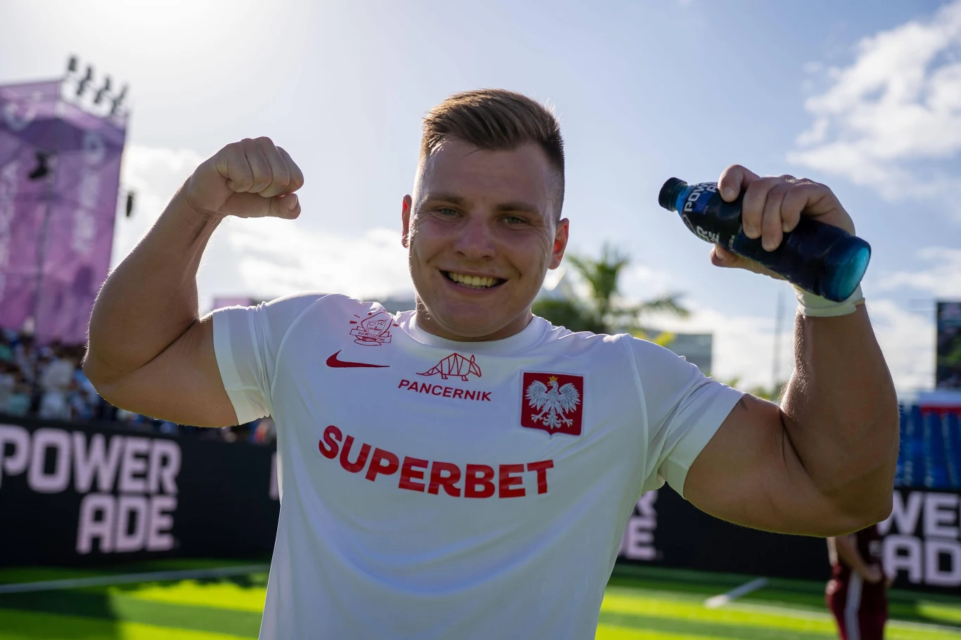 Norbert Jaszczak podczas mistrzostw świata w Socca Sportowiec w białej koszulce z orłem i napisem 'SUPERBET', unoszący ramiona w geście siły, uśmiechnięty, na tle stadionu i nieba z palmami.