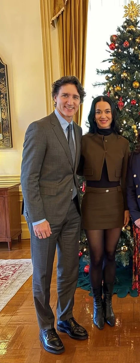 Katy Perry i Justin Trudeau na spotkaniu z byłym premierem Japonii i jego żoną Mężczyzna w szarym garniturze i kobieta w brązowym kostiumie stoją razem uśmiechnięci obok udekorowanej choinki w eleganckim, jasnym pomieszczeniu.