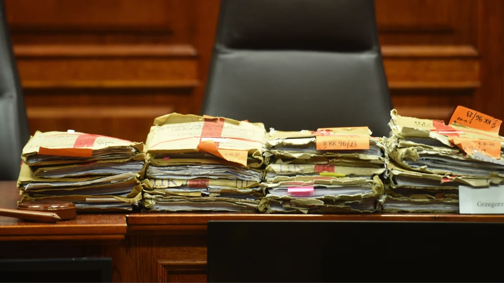 Stosy akt i dokumentów w papierowych teczkach ułożone na drewnianym stole, na tle pustego fotela w sali sądowej. Obok stosów znajduje się sędziowski młotek.