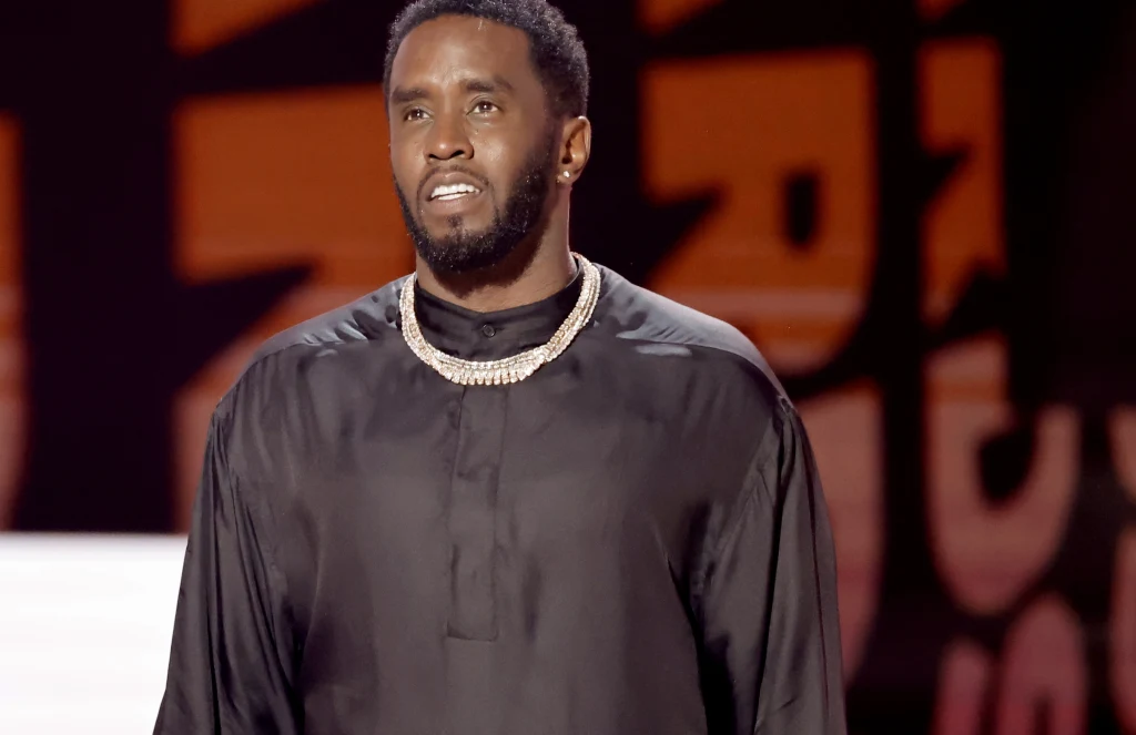 Czarnoskóry mężczyzna z krótkimi włosami i brodą ubrany w czarną koszulę oraz wyrazisty łańcuch na szyi stoi na tle graficznego napisu, patrząc w nieokreślonym kierunku.