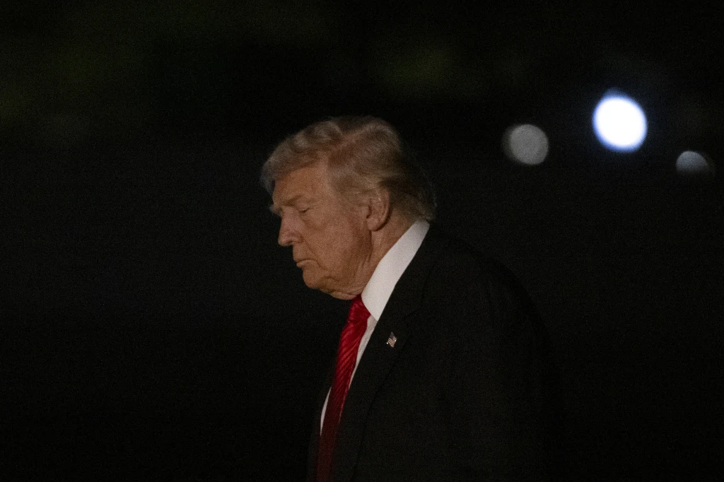 Prezydent USA Donald Trump Starszy mężczyzna w garniturze i czerwonym krawacie na ciemnym tle, światła w oddali, wyraz twarzy poważny, sylwetka z profilu.