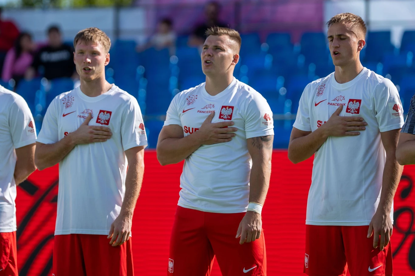 Reprezentacja Polski na mistrzostwach świata w Socca Trzech sportowców ubranych w białe koszulki z herbem Polski i czerwone spodenki, stojących z ręką na sercu podczas hymnu narodowego, w tle widać niebieskie trybuny stadionu.