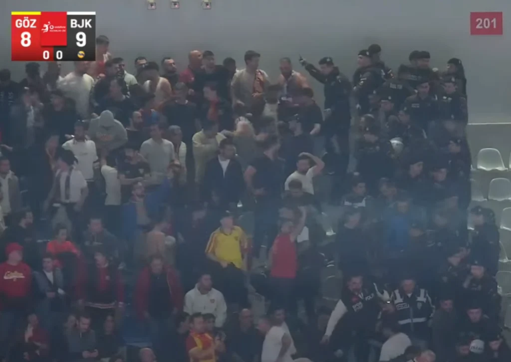 Tłum kibiców sportowych wypełniających trybuny stadionu, niektórzy stojący, unoszący ręce lub skandujący, z widocznymi elementami dymu i pustymi krzesłami po prawej stronie. W lewym górnym rogu widoczne wyniki meczu.