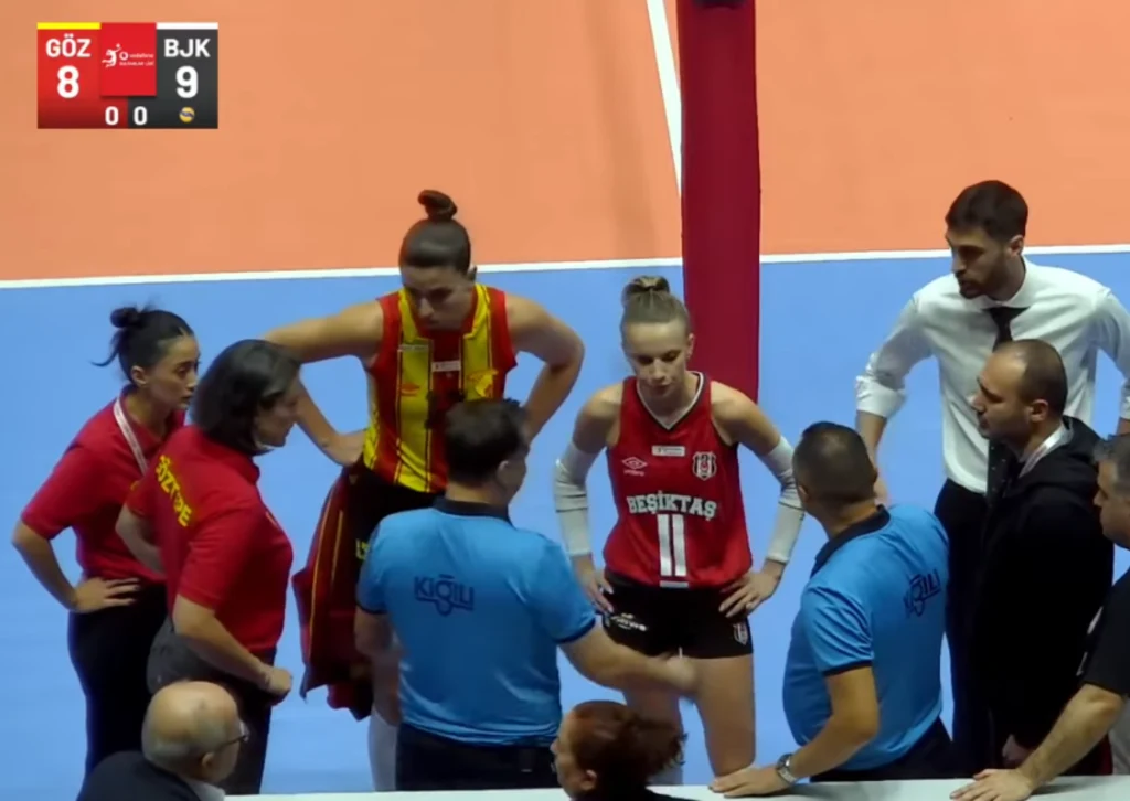 Grupa zawodniczek i trenerów obu drużyn rozmawia z sędziami przy stole sędziowskim podczas przerwy technicznej w meczu siatkówki na hali sportowej. Na planszy wyników widoczny aktualny wynik spotkania.