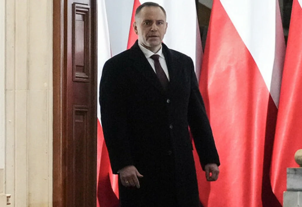 Prezydent Karol Nawrocki Mężczyzna w garniturze i płaszczu stoi na tle kilku dużych biało-czerwonych flag narodowych. Wnętrze budynku o oficjalnym charakterze, widoczne drzwi i fragment marmurowej kolumny.