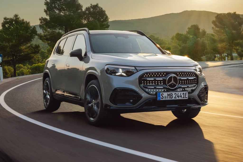 Nowoczesny srebrny SUV marki Mercedes-Benz na zakręcie asfaltowej drogi, oświetlony promieniami zachodzącego słońca. W tle drzewa i wzgórza tworzące spokojny krajobraz.