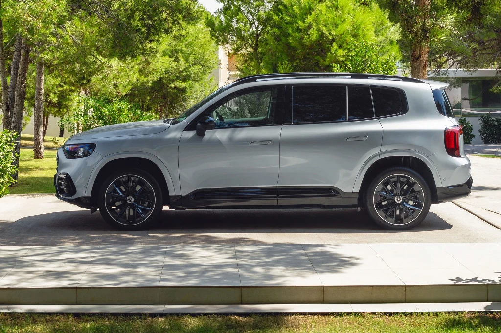 Srebrny SUV zaparkowany na podjeździe wśród zielonych drzew, z nowoczesnymi felgami i sportową sylwetką, częściowo w cieniu, na tle współczesnego budynku.