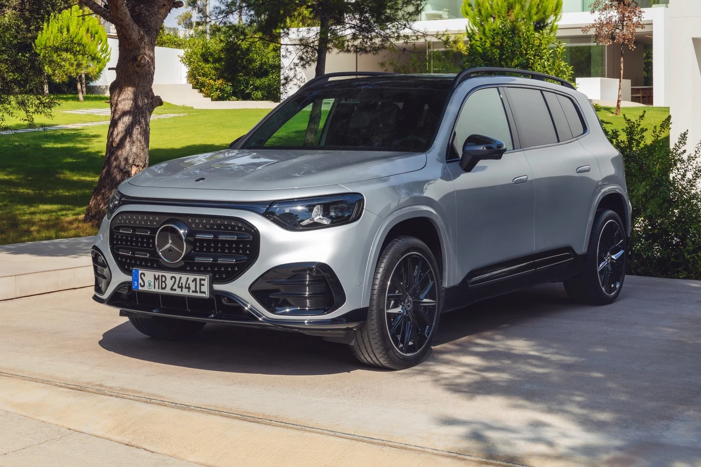 Srebrny SUV marki Mercedes-Benz stoi zaparkowany na podjeździe przed nowoczesnym budynkiem otoczonym zielenią, auto posiada czarne felgi i wyrazisty grill, światło dzienne podkreśla elegancką sylwetkę samochodu.