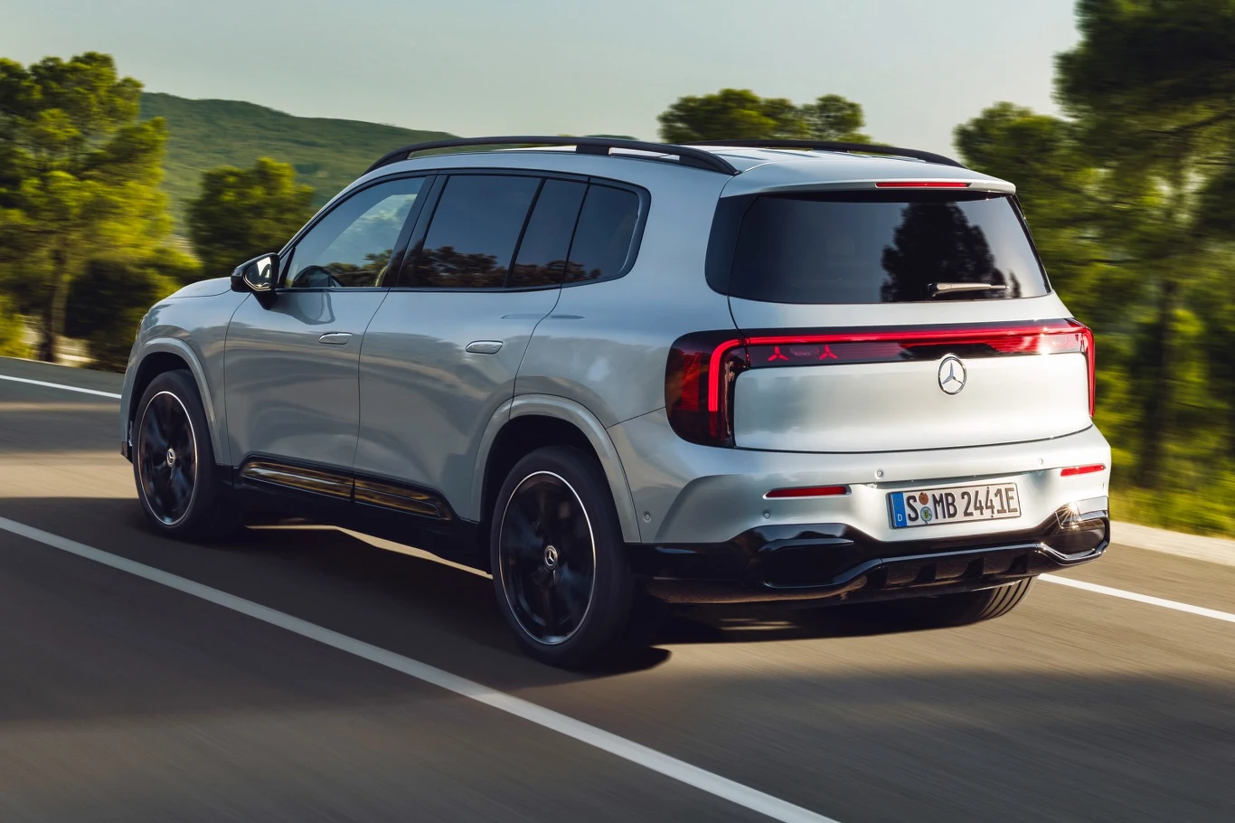 Srebrny, nowoczesny SUV marki Mercedes-Benz porusza się asfaltową drogą otoczoną zielenią i wzgórzami, auto wyróżnia się panoramicznym dachem, przyciemnianymi szybami oraz stylowymi felgami.