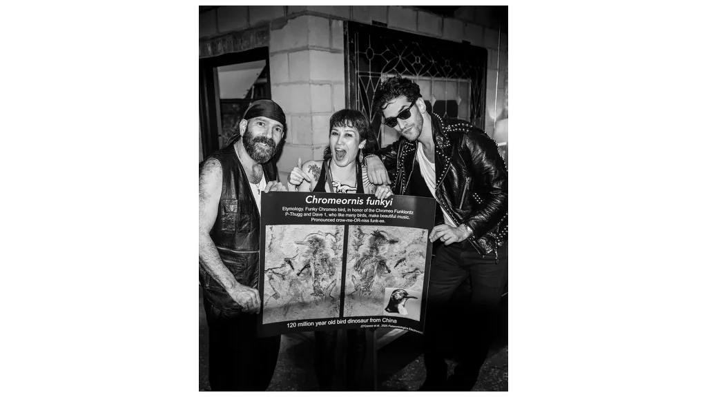 Trójka młodych dorosłych pozuje razem z plakatem przyklejonym do deski, ubrani w skórzane kurtki i ciemne okulary, wyrażając radość i zabawę, na tle miejskiego budynku.