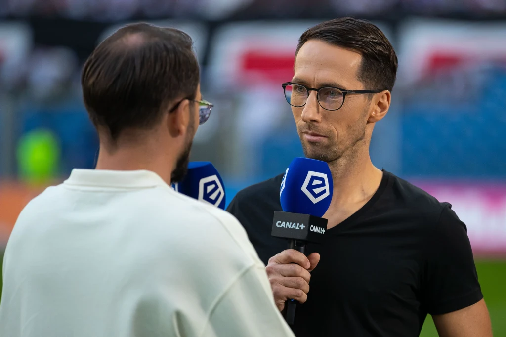Marcin Kikut Dwóch mężczyzn podczas wywiadu na stadionie, trzymających mikrofony z logo Canal+ oraz Ekstraklasy. Jeden z nich zwrócony przodem do obiektywu, drugi tyłem. W tle rozmyta trybuna stadionu.