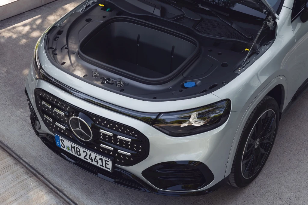 Otwarta maska białego samochodu elektrycznego marki Mercedes-Benz z widocznym przednim bagażnikiem. Wokół pojazdu znajduje się betonowe podłoże, a auto stoi zaparkowane, podkreślając nowoczesny charakter oraz design przodu samochodu.