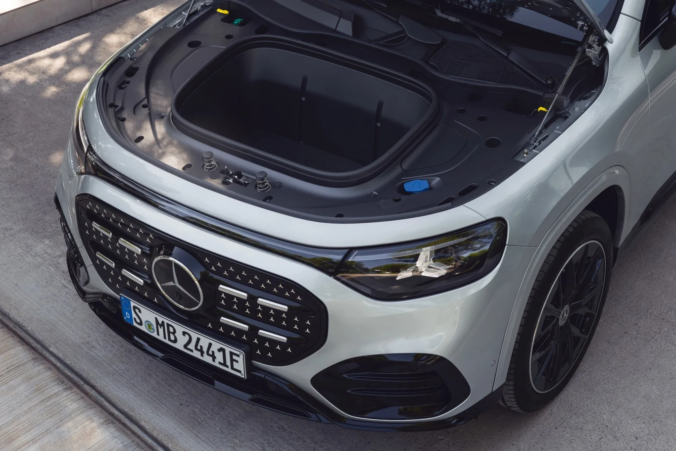 Otwarta maska białego samochodu elektrycznego marki Mercedes-Benz z widocznym przednim bagażnikiem. Wokół pojazdu znajduje się betonowe podłoże, a auto stoi zaparkowane, podkreślając nowoczesny charakter oraz design przodu samochodu.