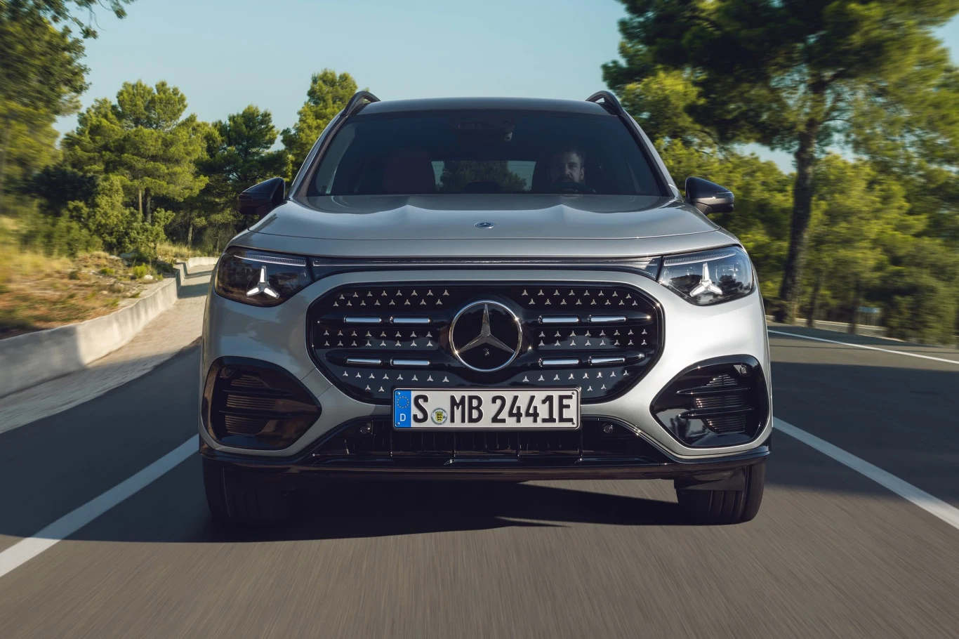 Srebrny samochód marki Mercedes z widocznym podświetlanym grillem i logo na środku maski, dynamicznie jadący po drodze otoczonej zielonymi drzewami w słoneczny dzień.