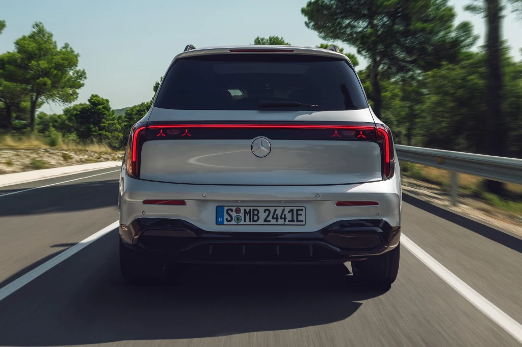 Luksusowy srebrny samochód osobowy marki Mercedes jedzie po równej asfaltowej drodze otoczonej zielonymi drzewami, widok z tyłu, na tablicy rejestracyjnej widoczny napis S MB 2441E.