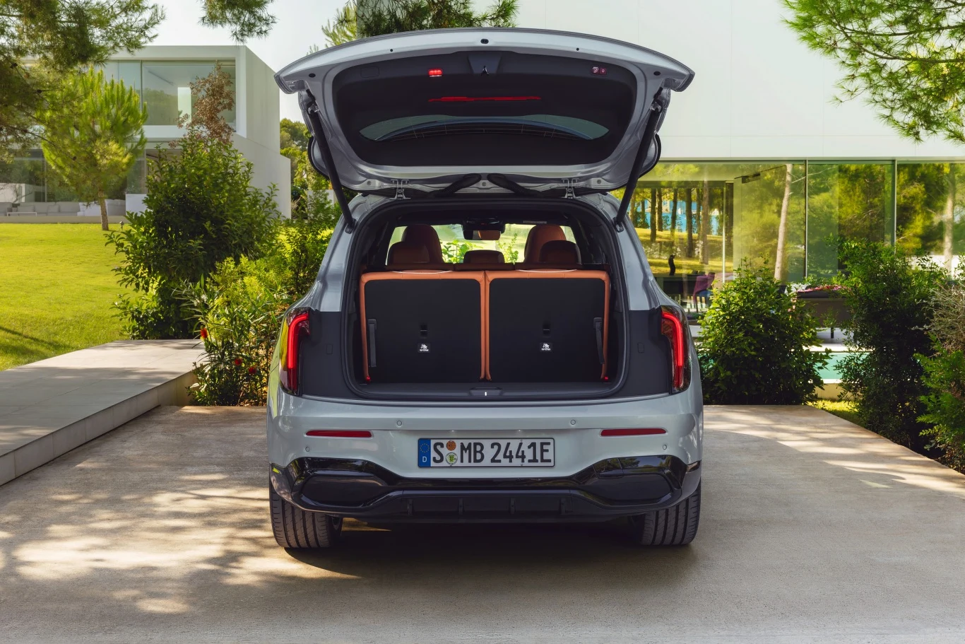 Otwarte bagażnik srebrnego samochodu osobowego zaparkowanego na podjeździe, w tle nowoczesny przeszklony budynek otoczony zielenią i drzewami.