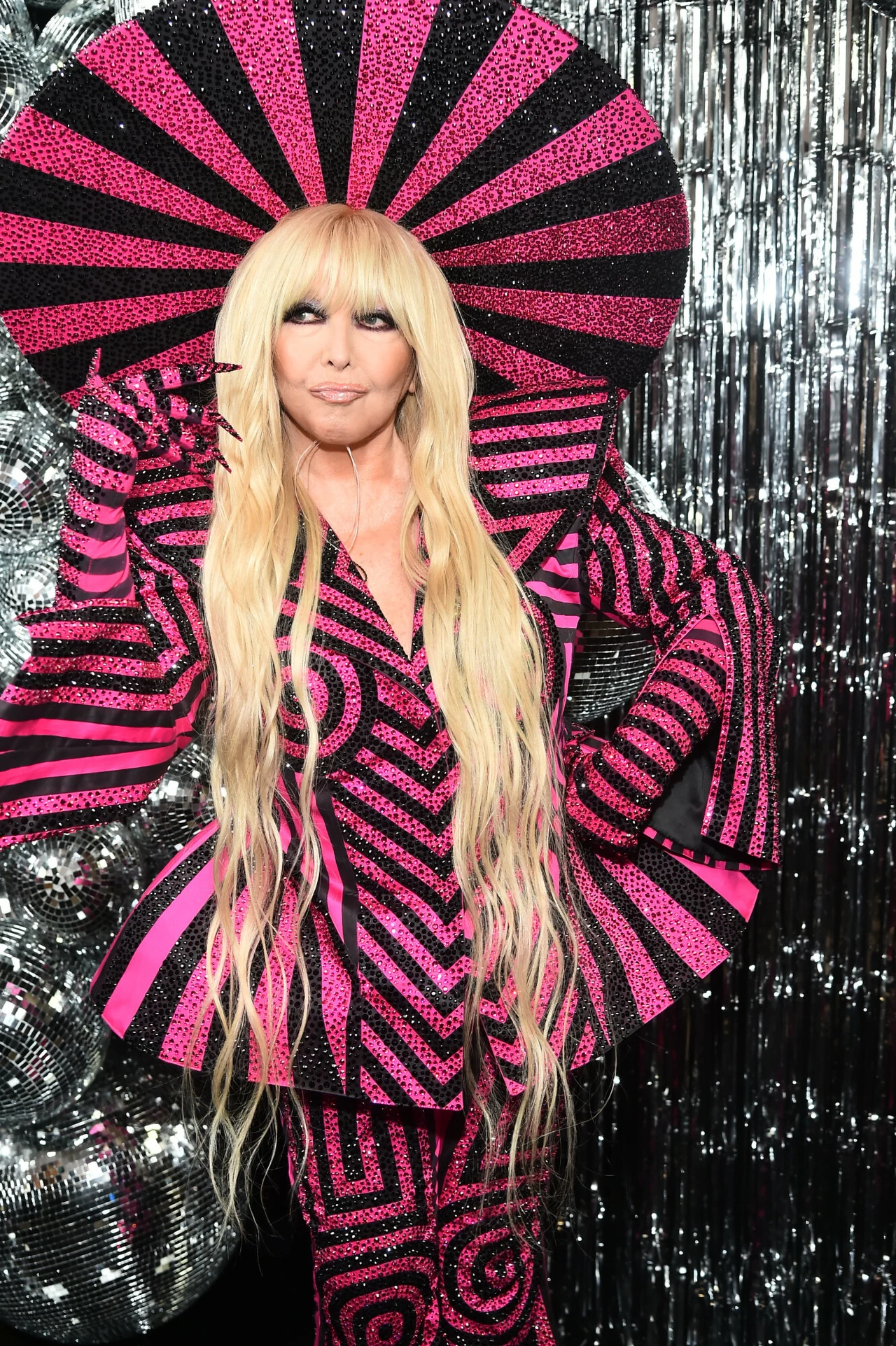 Maryla Rodowicz. Sylwestrowa Moc Przebojów w Toruniu 2024 Osoba w stroju sceniczno-artystycznym o wyrazistym wzorze w czarno-różowe paski i geometryczne kształty, z efektownym dużym nakryciem głowy w tym samym stylu, długie blond włosy rozpuszczone, tło ze srebrzystymi dekoracjami przypominającymi dyskotekowe...