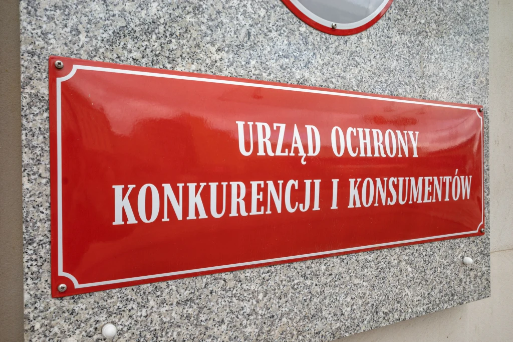 Czerwona tablica informacyjna z białym napisem Urząd Ochrony Konkurencji i Konsumentów, zamontowana na ścianie z szarego kamienia.